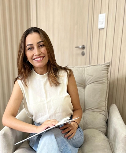 Antônia Honorato, sorrindo, de camisa sem manga bege e calça jeans, sentada em um sofá, com uma prancheta no colo